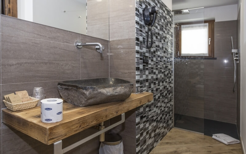 Modern bathroom with a stone sink, wooden countertop, glass shower, and mosaic-tiled walls, exuding cleanliness and sophistication.