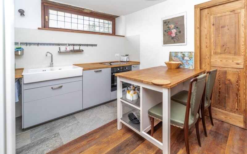 Bright kitchen with white walls, wooden accents, farmhouse sink, gray cabinets, and a cozy wooden table with green chairs.