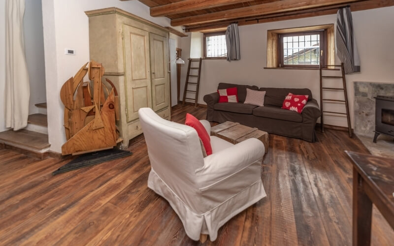 Cozy rustic living room with wooden flooring, brown couch, white armchair, wooden crate table, and a black wood-burning stove.