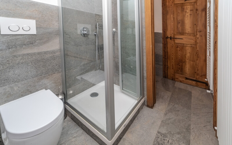 A modern bathroom featuring a glass shower, white toilet, gray tiled walls, and a wooden door with black handles.