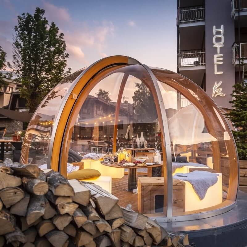 Transparent dome with wooden frame in front of a hotel, featuring two beds, a table, and firewood, surrounded by trees.