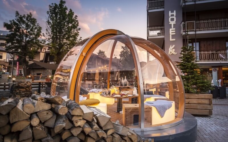 Transparent dome with wooden interior, two beds, dining table, Christmas tree, and hotel in the background under a pinkish-blue sky.