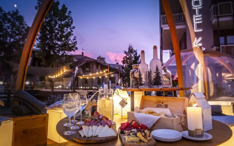 A round wooden table set with food and drinks, against a hotel backdrop with trees and a sunset sky.