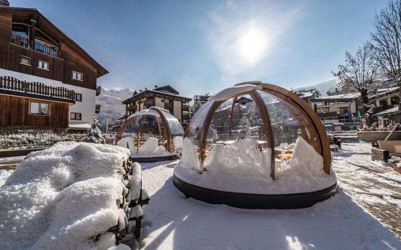 Due grandi strutture a cupola trasparenti in una zona innevata, circondate da alberi e edifici, sotto un cielo sereno.