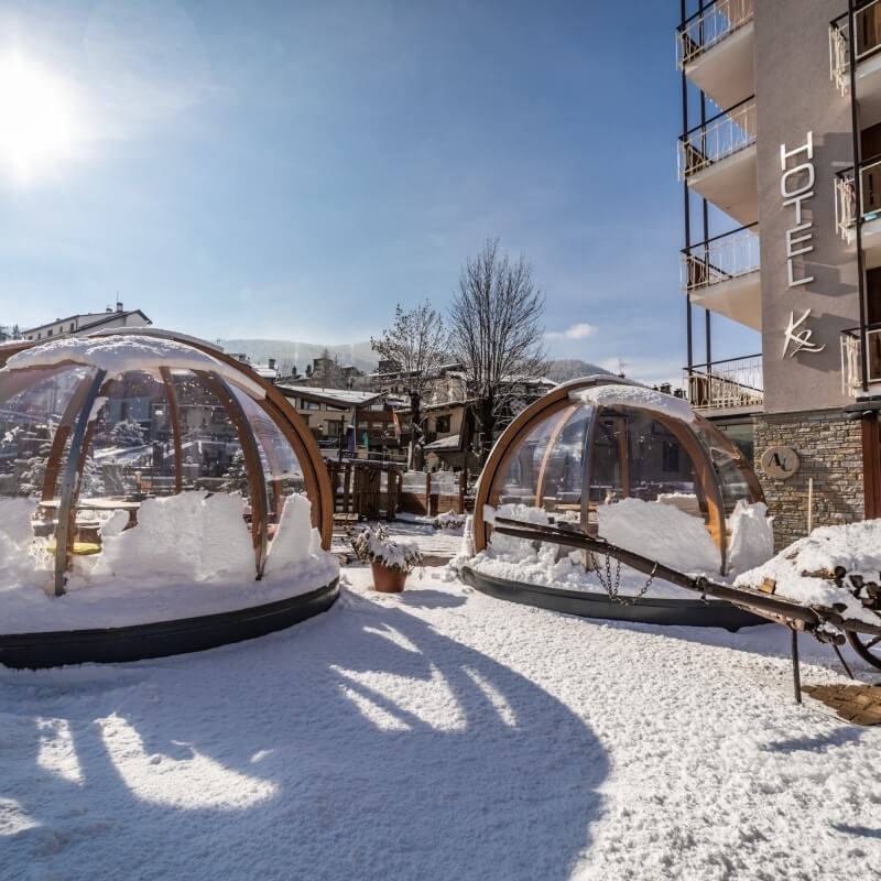 Snowy scene with two transparent domes by a hotel, a "HOTEL" sign, and a clear blue sky on a winter day.