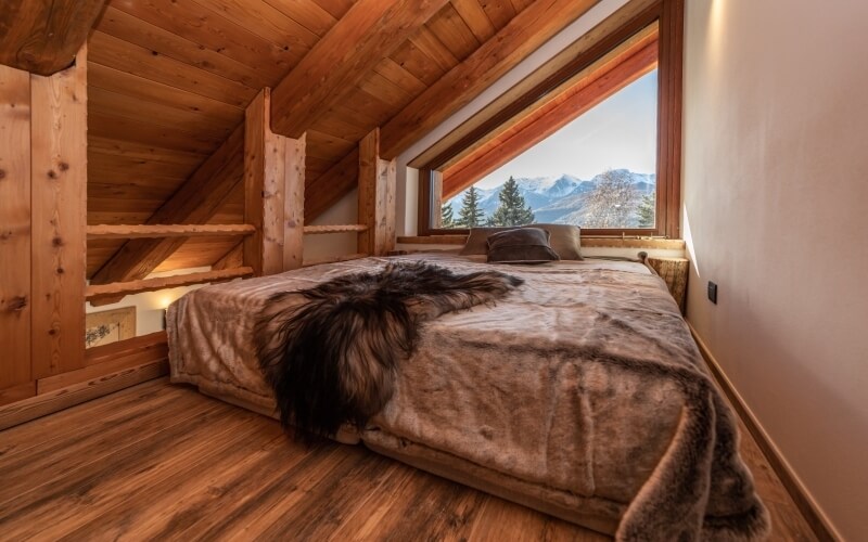Cozy bedroom with wooden architecture, large window view of mountains, inviting bed with brown blanket and pillows.