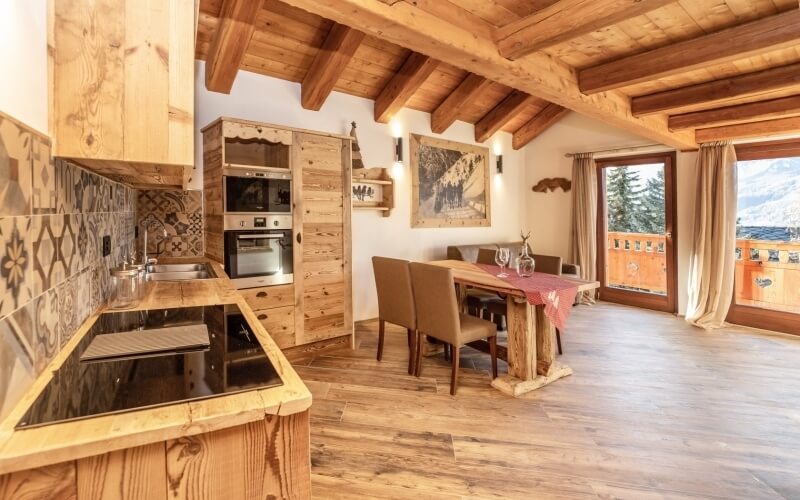 Cucina rustica con isola in legno, tavolo con tovaglia rossa e vista su alberi e montagne attraverso grandi finestre.