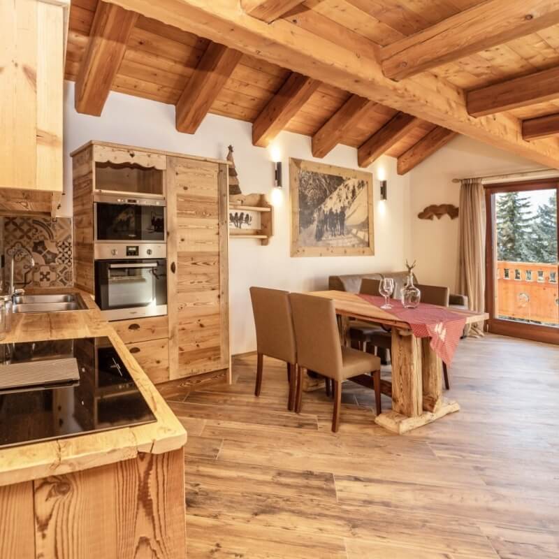 Rustic open-plan kitchen and dining area with wooden beams, a red tablecloth, wine glasses, and a view of trees outside.