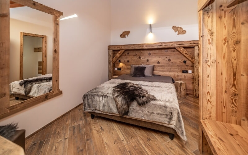 Cozy rustic bedroom with a wooden bed, mirror, gray comforter, and white fur blanket, featuring wooden floors and walls.