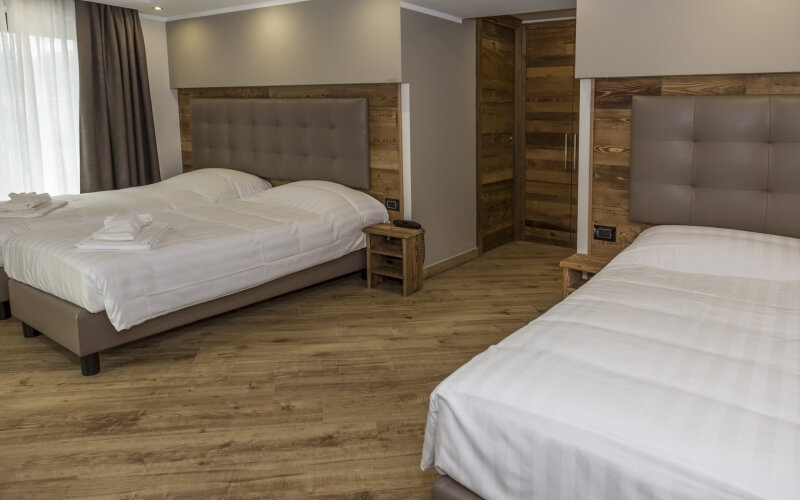 Hotel room with a double bed and single bed, white comforters, wooden table, light brown walls, and natural light from a window.