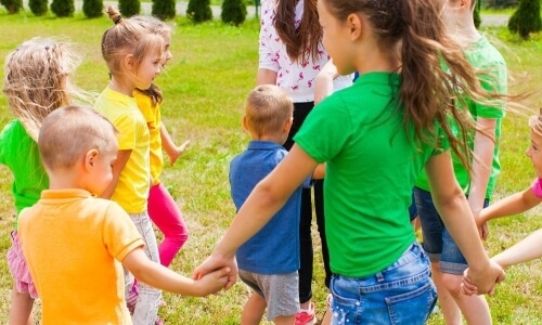 A group of children and adults holding hands in a circle, set in a grassy area, suggesting a summer camp activity.