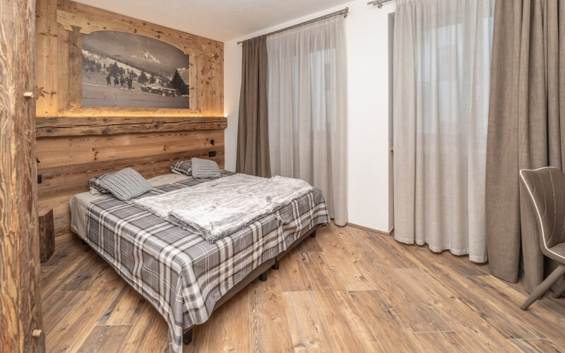 Cozy rustic bedroom with a plaid comforter, wooden headboard, chair, large winter painting, and sheer-curtained windows.