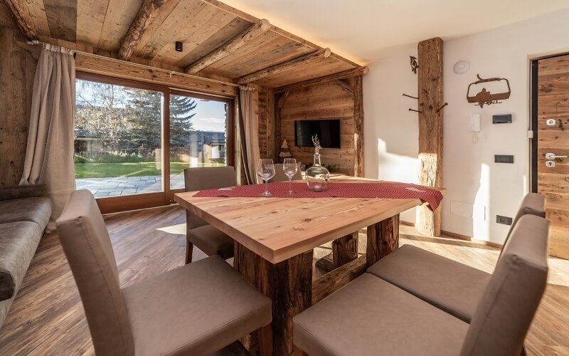 Rustic open-plan living area with wooden elements, dining table, gray couch, and serene outdoor view through a large window.