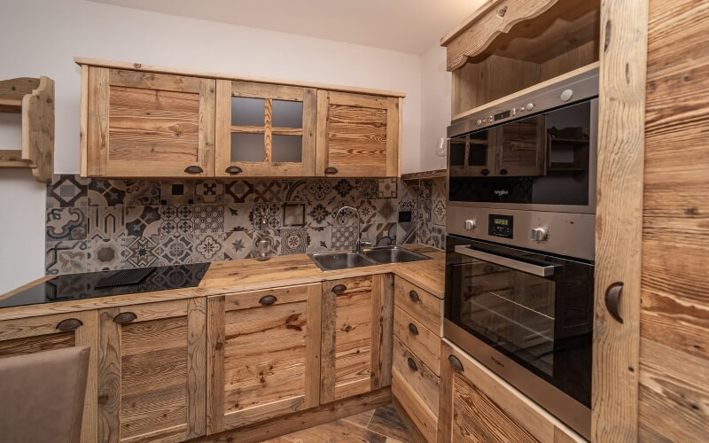 A cozy kitchen with wooden cabinets, patterned tile backsplash, built-in oven, stainless steel sink, and slanted ceiling.