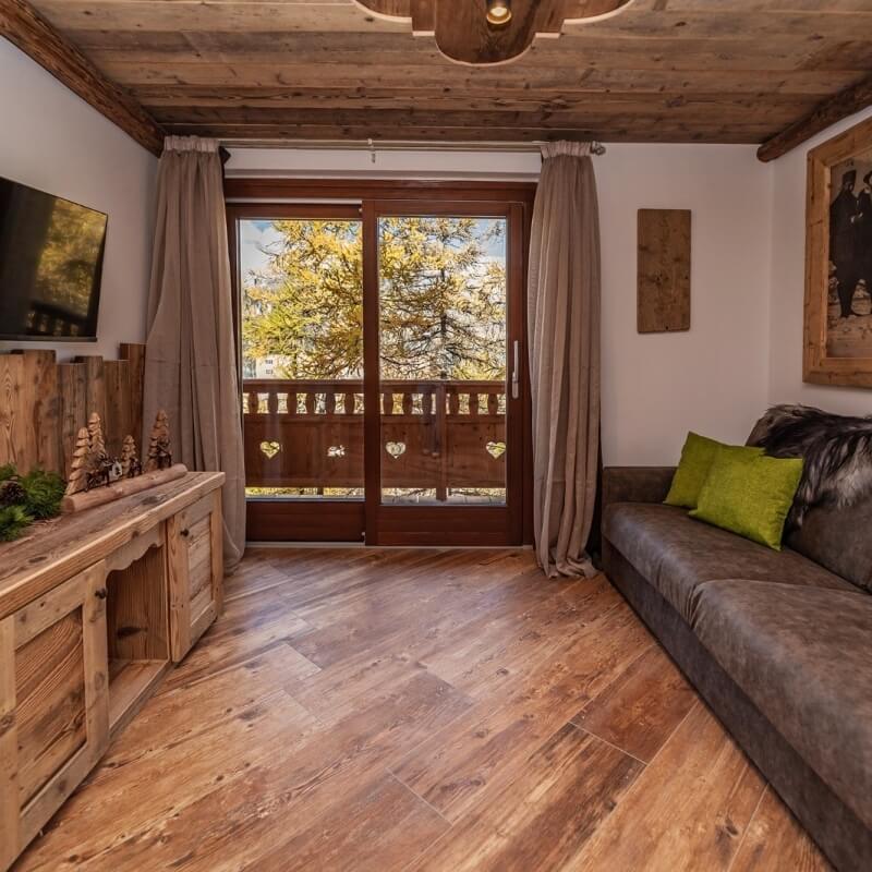 Cozy rustic living room with a fireplace, large window, dark gray sofa, and autumn view of yellow-leaved trees outside.