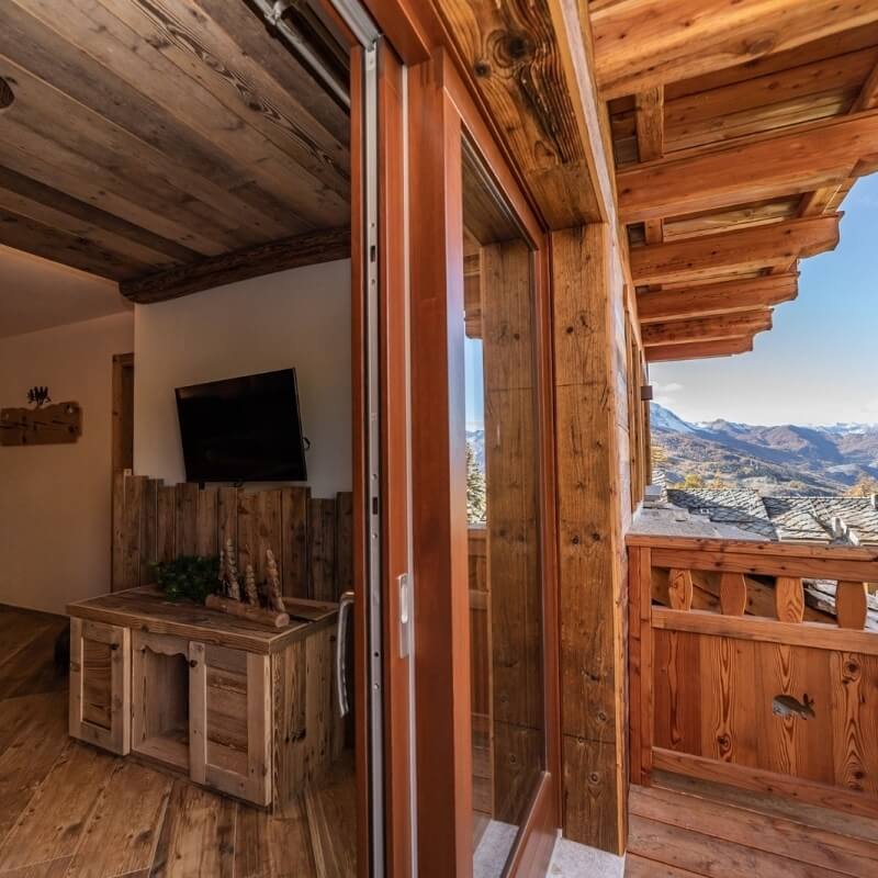 Wood-paneled room with a glass door to a balcony, showcasing mountain views, wooden floors, and a small cabinet with a TV.