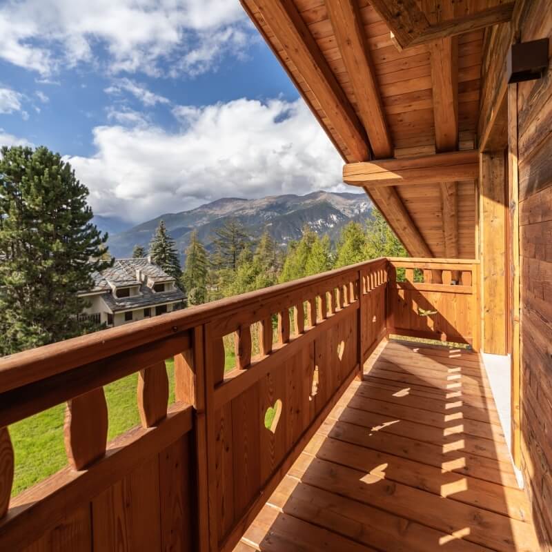 Balcone in legno con ringhiera a cuori, vista su alberi e montagne sullo sfondo. Casa bianca parzialmente visibile.