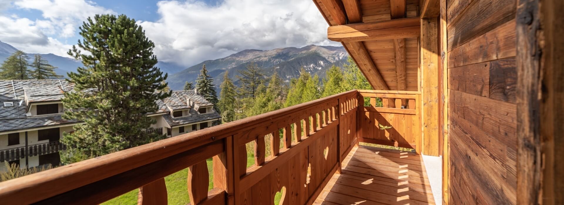 Wooden balcony with railing overlooking trees and mountains under a blue sky with clouds, creating a serene atmosphere.