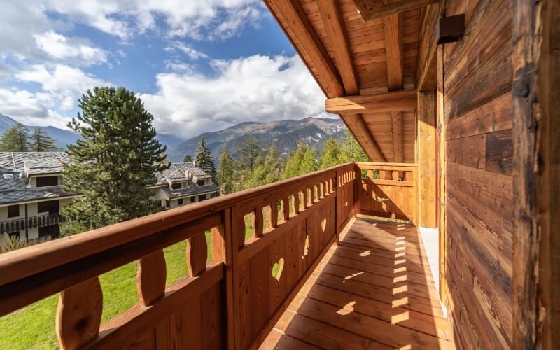 Wooden balcony overlooking majestic mountains, lush trees, and a blue sky with clouds, exuding tranquility and rustic charm.