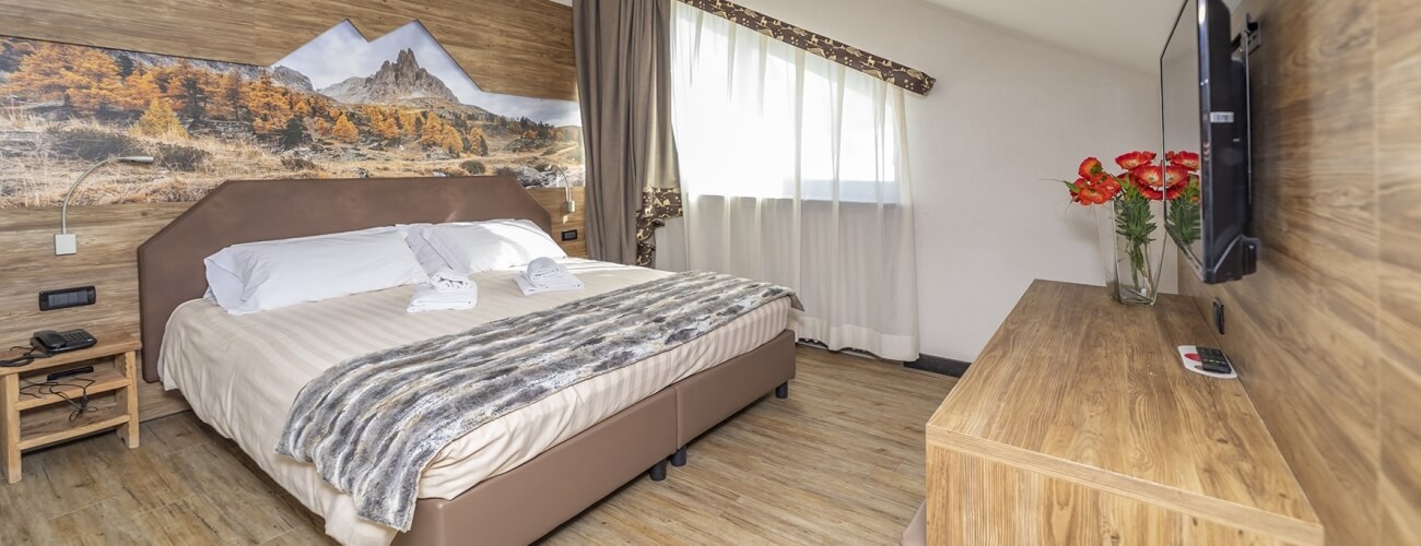 Camera d'hotel con letto grande, TV, decorazioni, e un murale di montagne dietro il letto. Pavimento in legno chiaro.