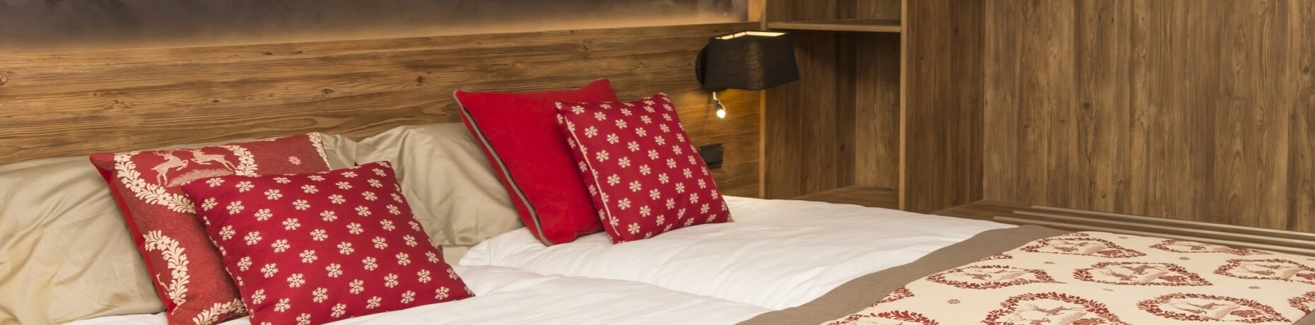 A bed with a wooden headboard, red and white pillows, and blankets, with a black lamp on a shelf against a dark brown wall.