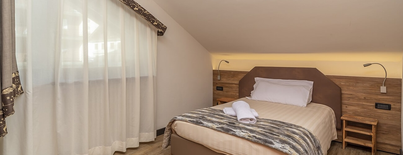 Serene bedroom with a brown headboard bed, tan comforter, white pillows, nightstand, lamp, and sheer curtains.