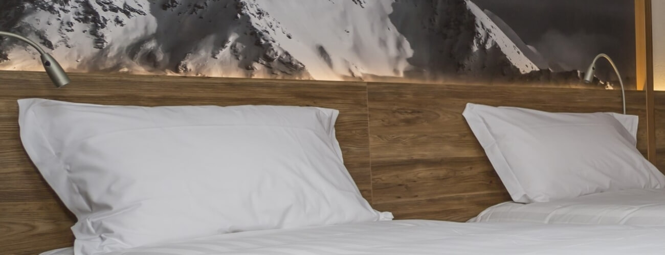 Letto con lenzuola bianche, testiera in legno, lampade calde e foto di montagne innevate sopra, creando un'atmosfera accogliente.