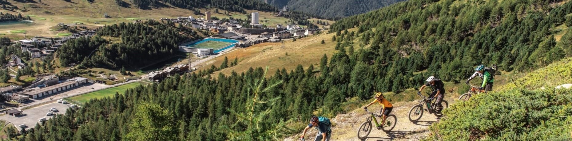 Ciclisti su un sentiero di montagna con un paese e una piscina sullo sfondo, circondati da alberi e natura.