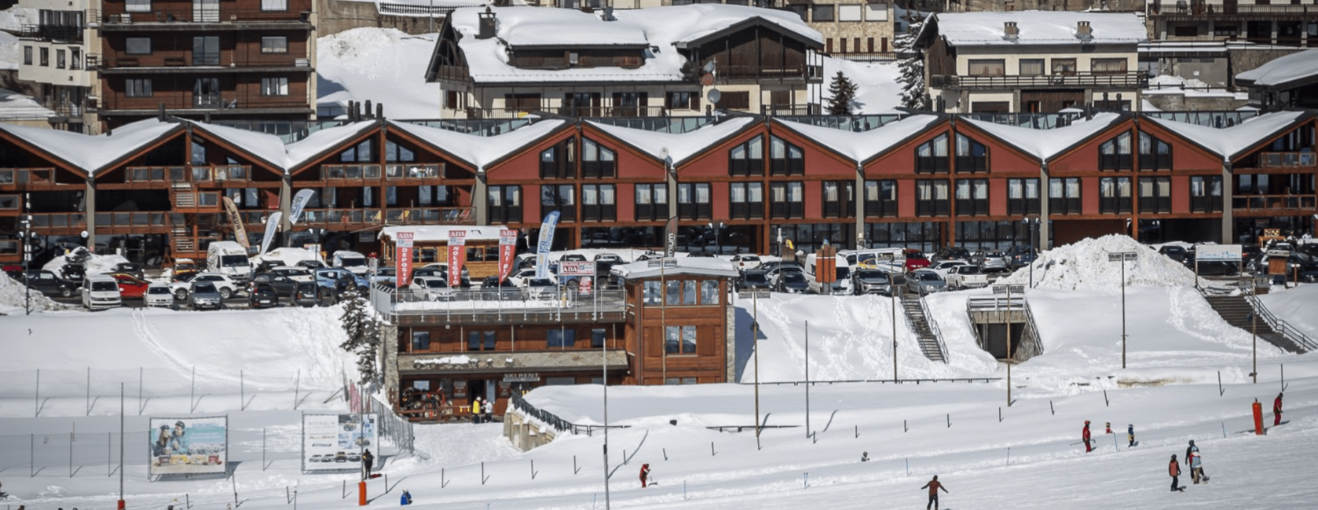 Snow-covered village with ski area, apartment buildings, shops, and cars, capturing a lively winter resort atmosphere.