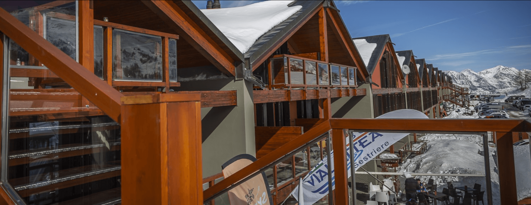 Paesaggio invernale con edifici a tetto spiovente, balconi in legno e montagne innevate sullo sfondo.