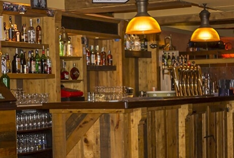 Cozy bar with wooden paneling, stocked shelves of beverages, warm lighting, and neatly arranged glasses in front.