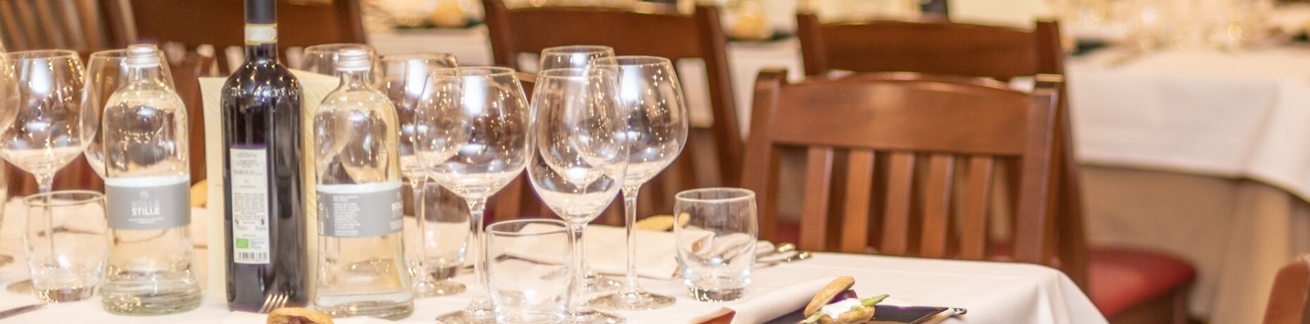 Elegant dining table set with a white tablecloth, various glassware, liquor bottles, and wooden chairs in the background.