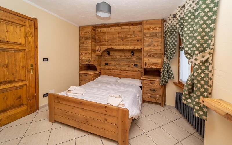 Rustic bedroom with a single bed, wooden furniture, green floral curtains, and beige tiled floor, exuding warmth and comfort.