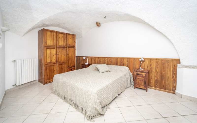 Camera da letto minimalista con pareti bianche, letto con coperta chiara, comodini, armadio e soffitto a volta.