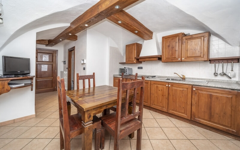 Cucina con area pranzo, mobili in legno, tavolo rettangolare, TV, grande isola con lavello e piano cottura elettrico.