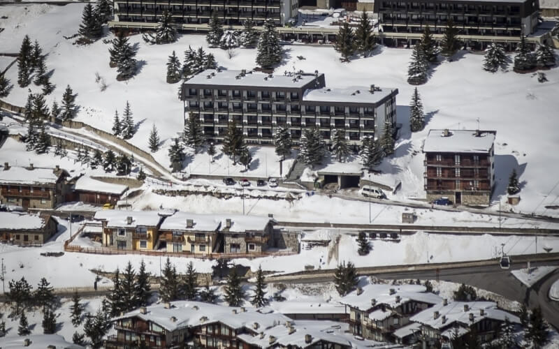 Aerial view of a snowy landscape with a large building, smaller structures, and trees, creating a serene winter scene.