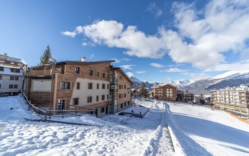 Resort di montagna innevato con edifici, una strada coperta di neve e montagne sullo sfondo.