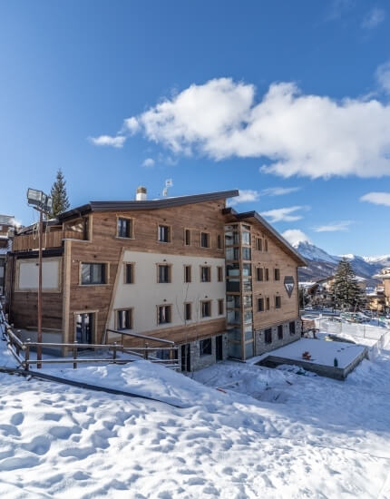 Wooden multi-story building in a snowy landscape, featuring a white facade, flat roof, and surrounded by mountains and trees.