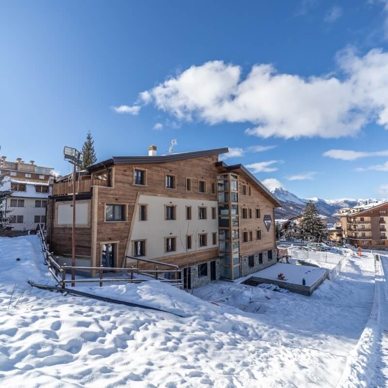 Edificio in legno a più piani con pareti bianche e marroni, circondato da neve e montagne sotto un cielo blu.