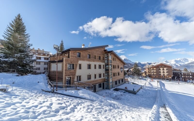 Serene winter scene with a wooden building on a snow-covered hill, blue sky, and distant trees, evoking tranquility.