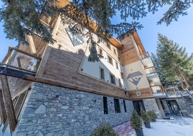 Low-angle view of a multi-story building with stone and wood, surrounded by snow-covered pine trees under a clear blue sky.