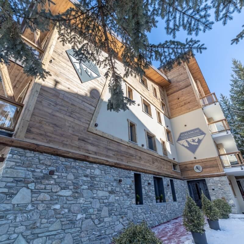 Edificio alpino a più piani con pietra grigia, legno, finestre, balconi e loghi "AVALANCHE" e "A" all'ingresso.