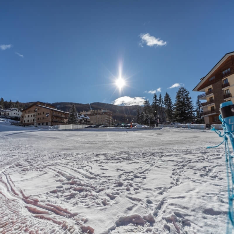 Scena invernale serena con edifici, montagne, neve e oggetti blu a destra. Cielo blu chiaro con alcune nuvole sparse.