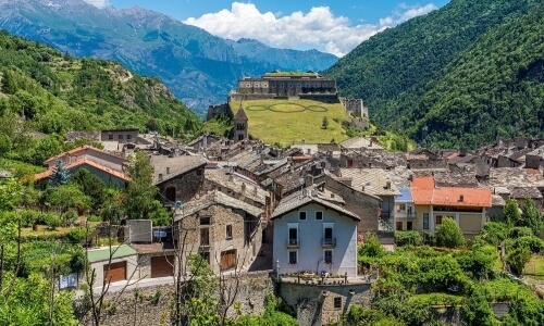 Villaggio di montagna sereno con case in pietra, fortezza sul colle, circondato da prati verdi e cielo blu.