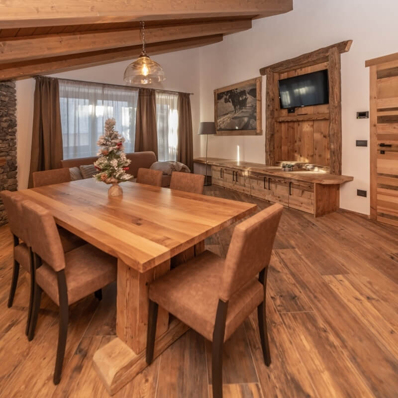 Cozy rustic living room with a dining area, wooden table, chairs, TV, and a small Christmas tree, featuring exposed beams.