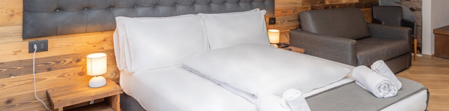 Cozy hotel room with a bed, nightstand, couch, and light wood wall; features white linens and a rolled towel on the floor.