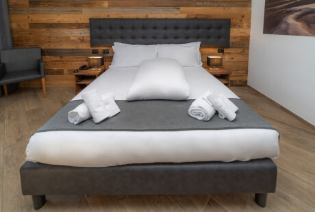 Serene bedroom with a gray tufted bed, white linens, wooden wall, nightstands with lamps, and a dark gray chair.