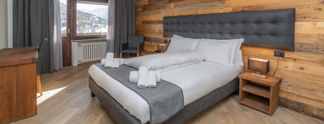 Camera d'albergo accogliente con letto grande, comodini, sedie nere, vista su montagne innevate e arredi in legno chiaro.