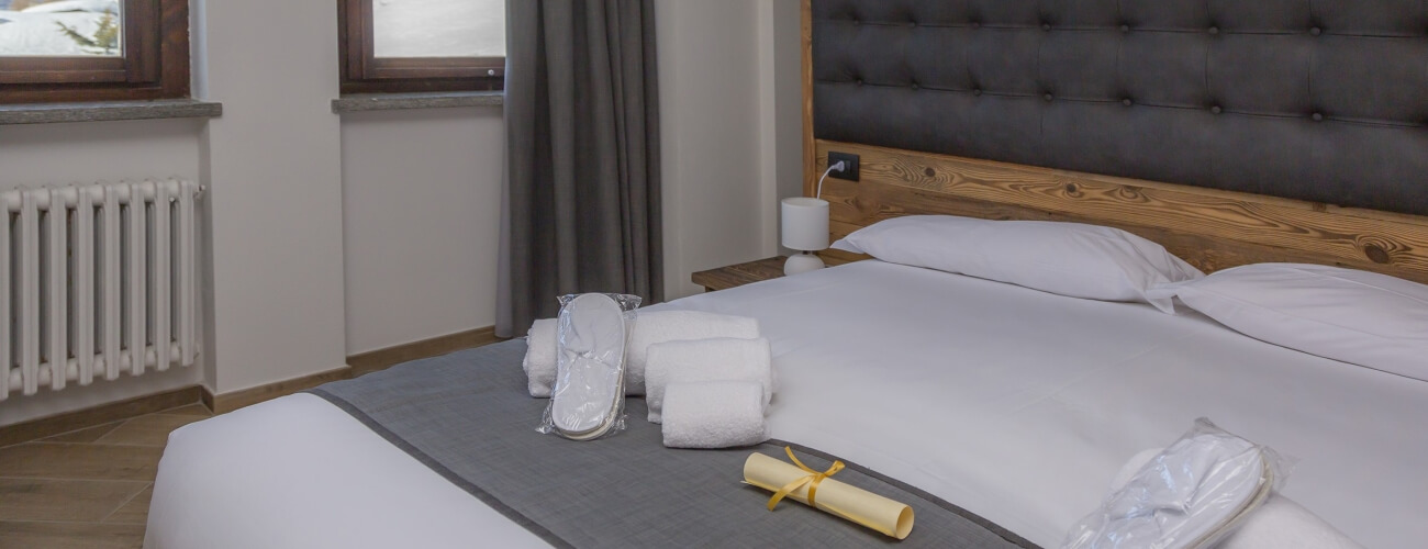 Hotel room featuring a made bed with white sheets, a scroll, slippers, towels, and a dark gray tufted headboard.