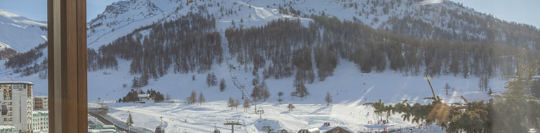 Serene winter landscape with snowy mountains, trees, ski lifts, and a wooden pillar from inside a ski lodge.